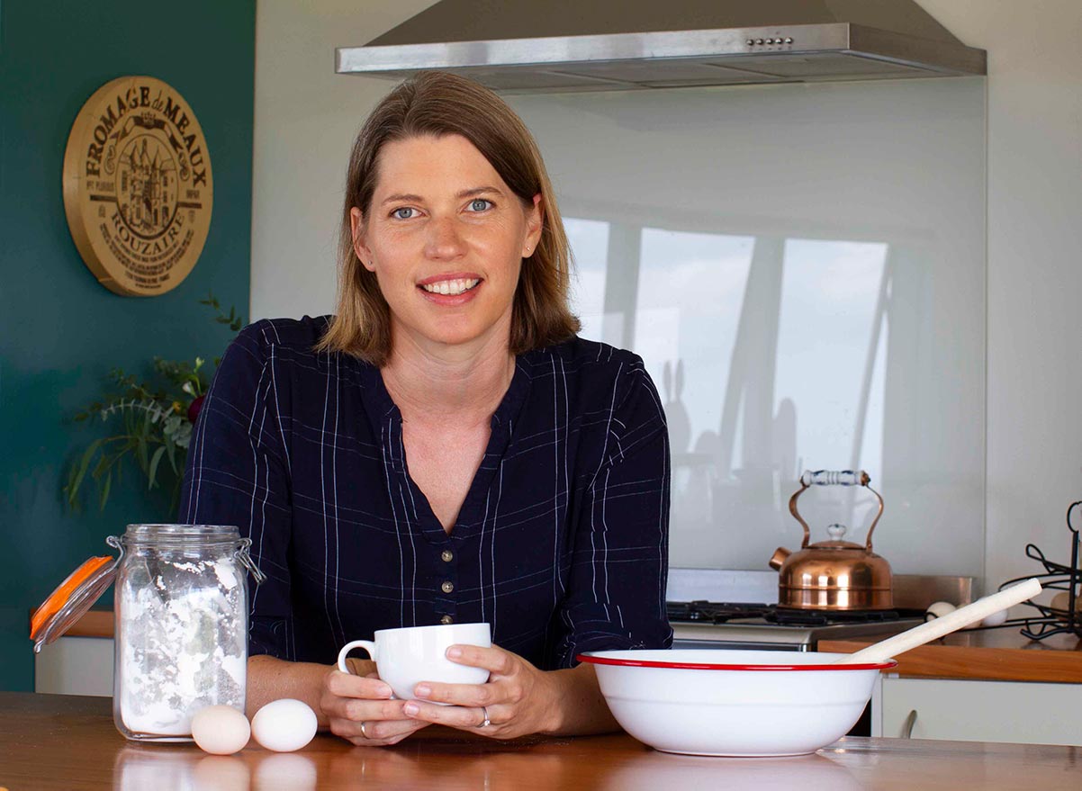 A woman with shoulder-length hair, wearing a dark plaid shirt, smiles while leaning on a kitchen counter. In front of her are a jar of flour, two eggs, and a white cup. A stove and kitchen utensils are in the background.