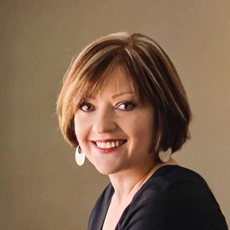 A woman with short, brown hair smiles at the camera. She is wearing large, circular earrings and a dark top. The background is simple and neutral-colored.
