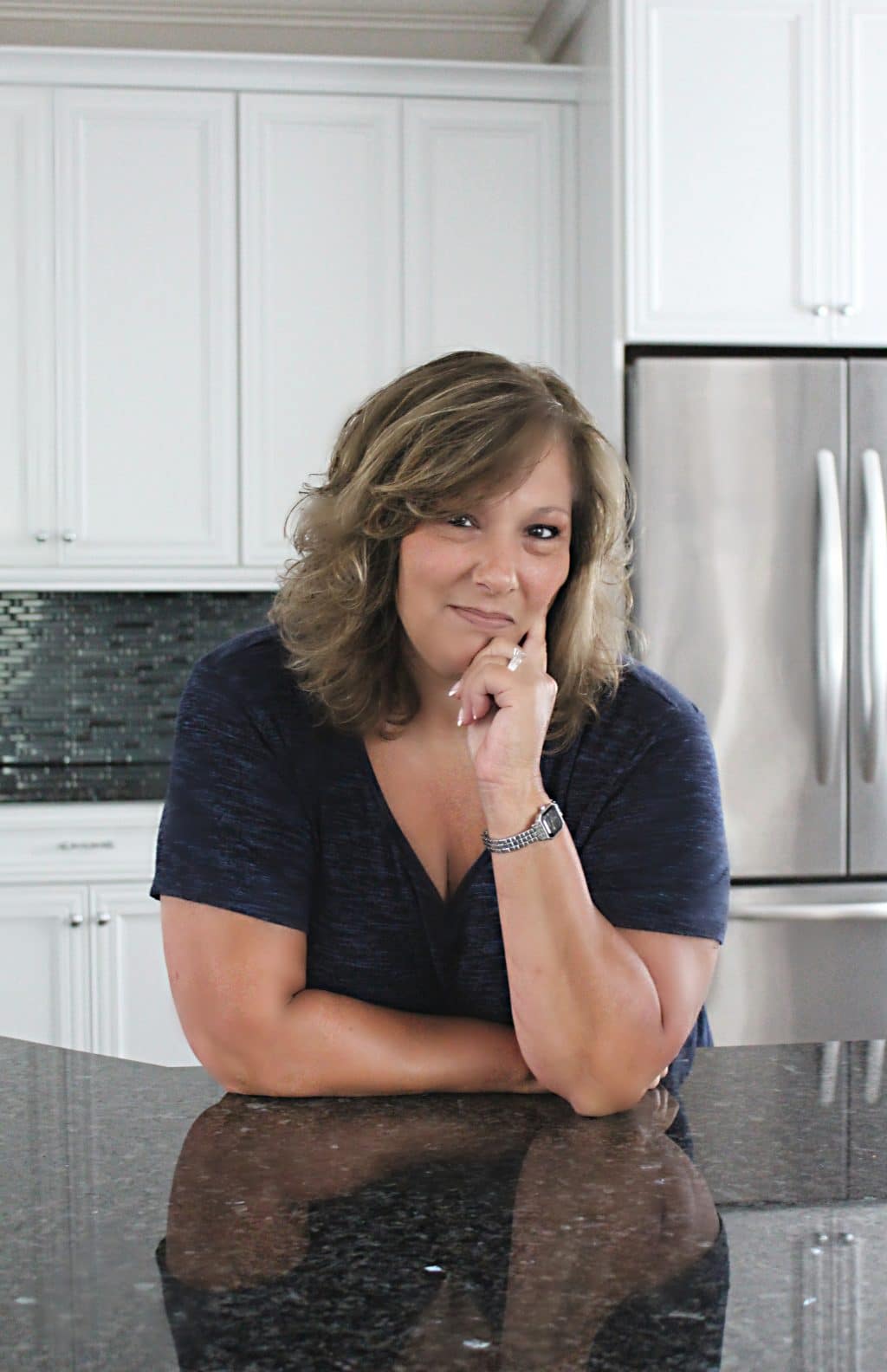 A person with wavy hair is leaning on a kitchen counter, resting their chin on their hand. They're wearing a dark blue shirt and a watch, with a neutral expression. The kitchen has white cabinets and a stainless steel refrigerator.