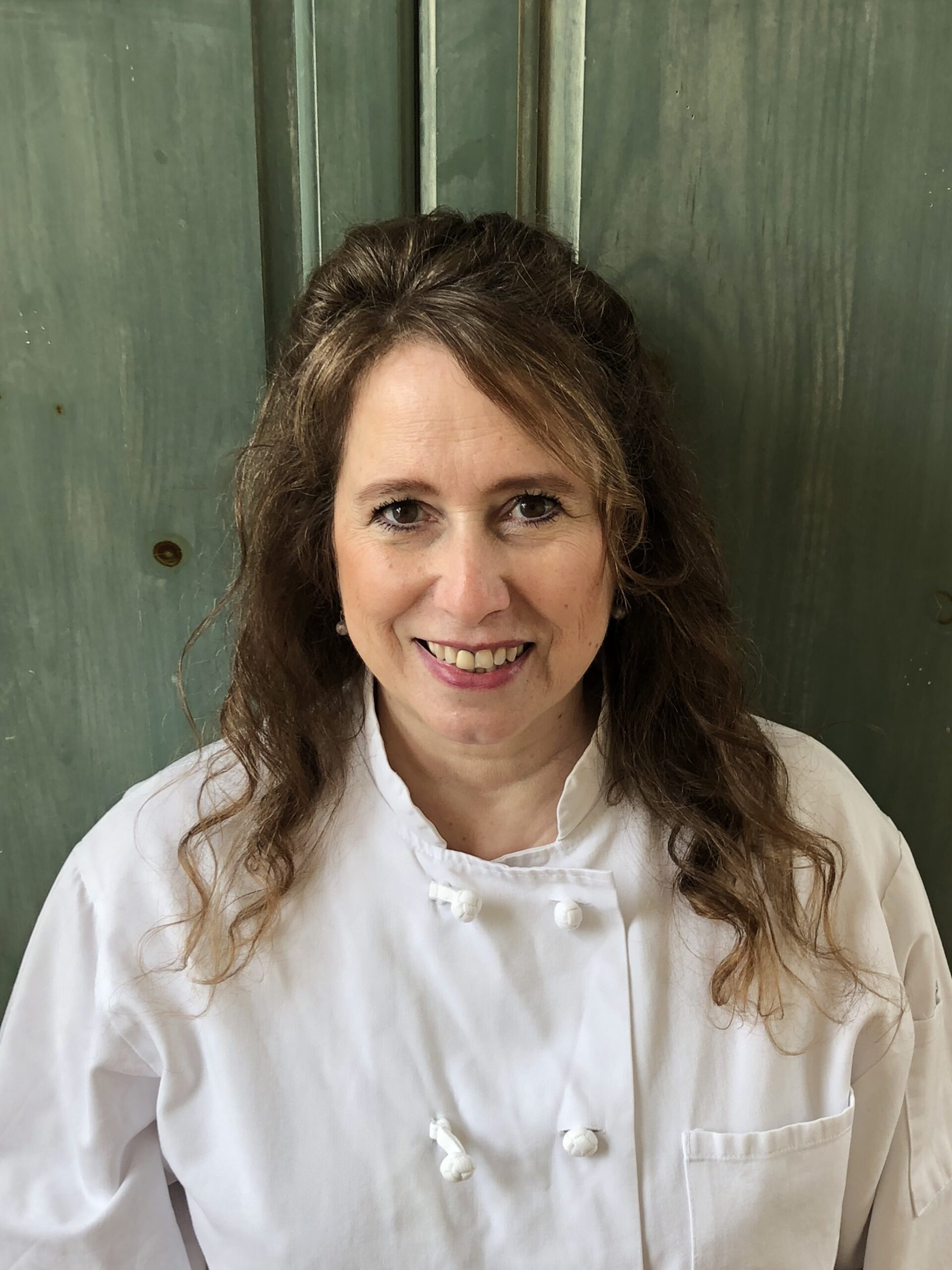 A smiling woman wearing a white chef's coat stands in front of a green wooden background. She has long, wavy brown hair and is looking directly at the camera.