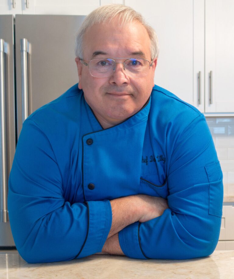 A man wearing glasses and a blue chef's coat leans on a kitchen counter with his arms crossed. The kitchen features white cabinets and a stainless steel refrigerator in the background.