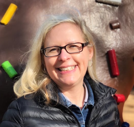 Smiling person with glasses and blonde hair, wearing a black jacket, stands outdoors in front of a background featuring colorful climbing holds.