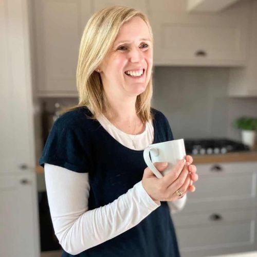 A person with blonde hair is smiling while holding a white mug in a kitchen. They are wearing a black shirt over a white long-sleeve top. The kitchen has light-colored cabinets and a stove in the background.