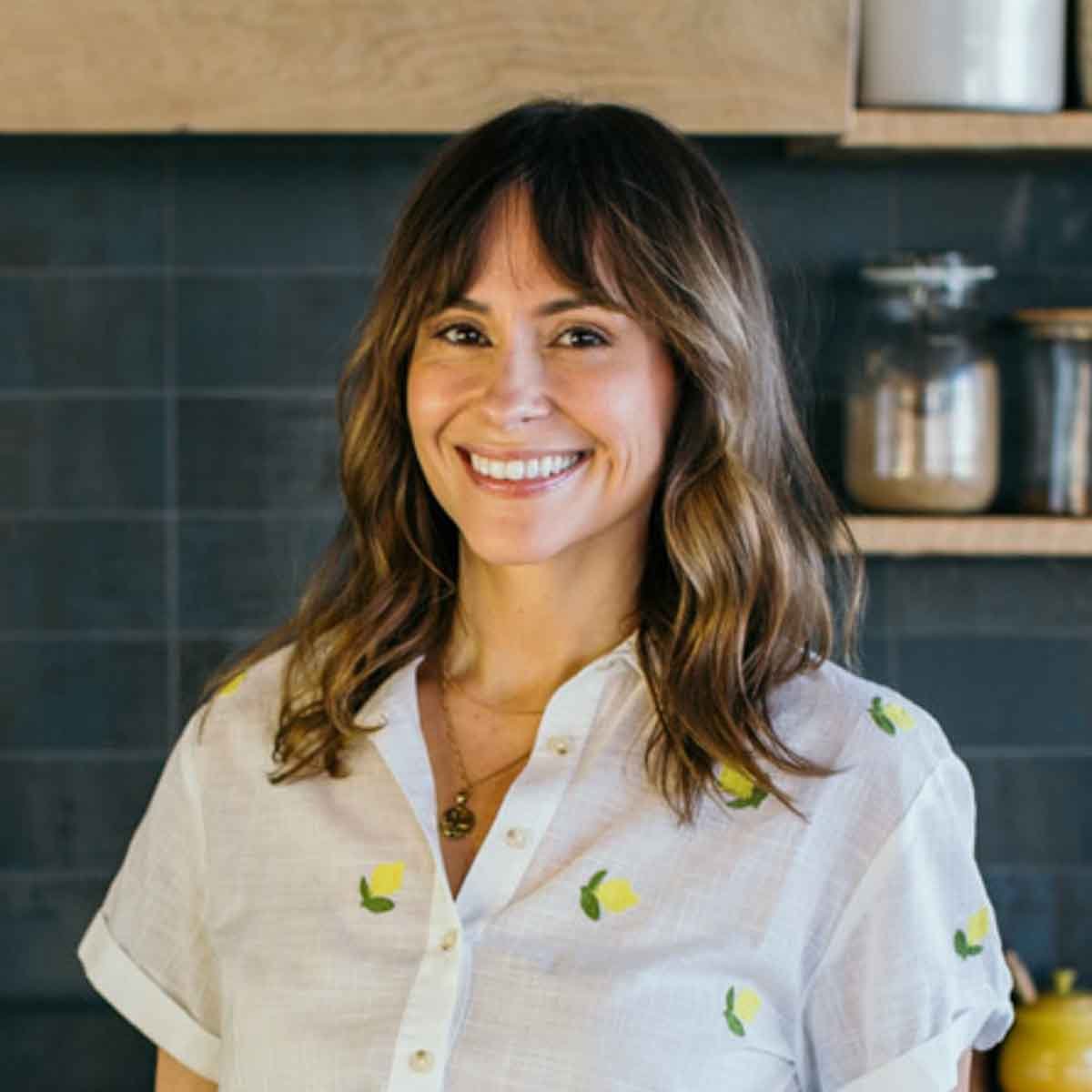 Smiling woman with shoulder-length brown hair stands in a kitchen, wearing a white shirt with yellow flower patterns. Dark tiled wall and shelves with jars are in the background.