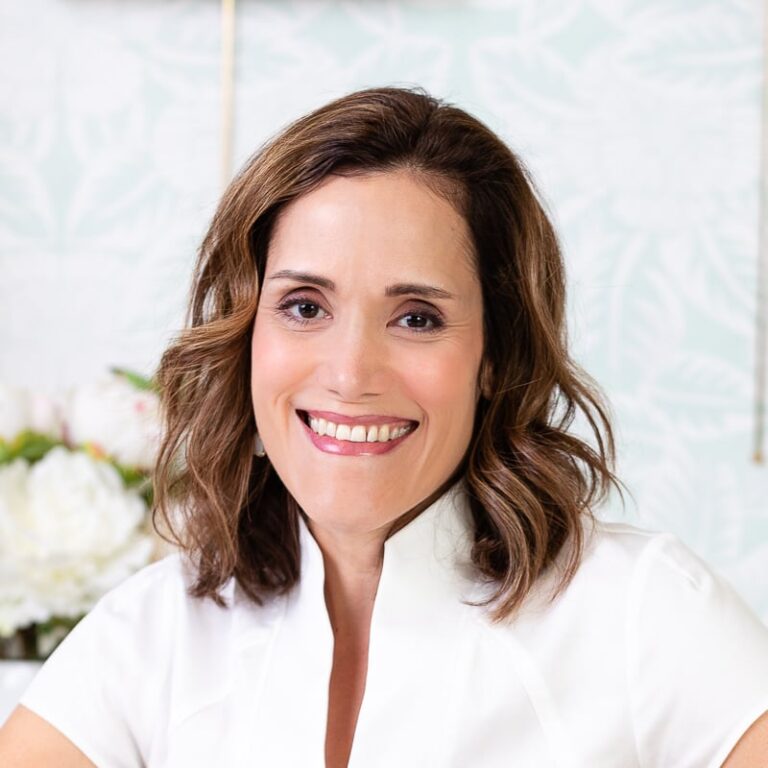 Smiling woman with shoulder-length brown hair wearing a white top, standing in front of a floral wallpaper background.