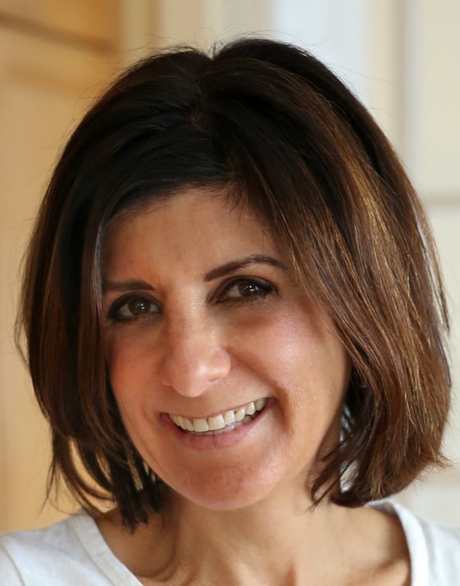Smiling person with short brown hair in a white top, standing indoors with a soft-focus background.