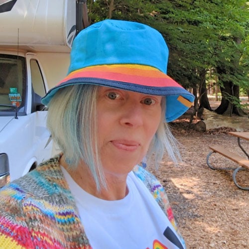 A person with light blue hair, wearing a blue bucket hat with a rainbow band and a colorful cardigan, stands outside near a camper van and a picnic table in a wooded area.