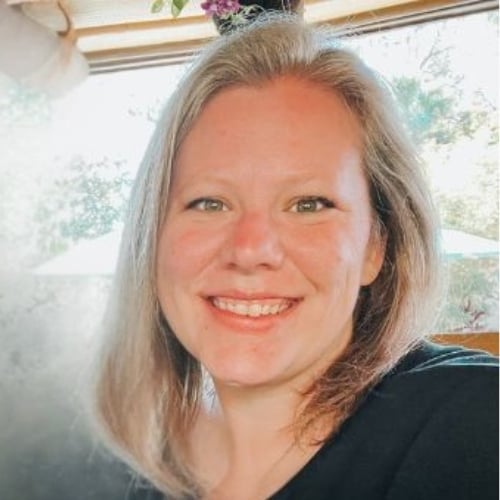 Smiling person with shoulder-length hair, wearing a black top, is seated in a brightly lit room with greenery visible through a window in the background.