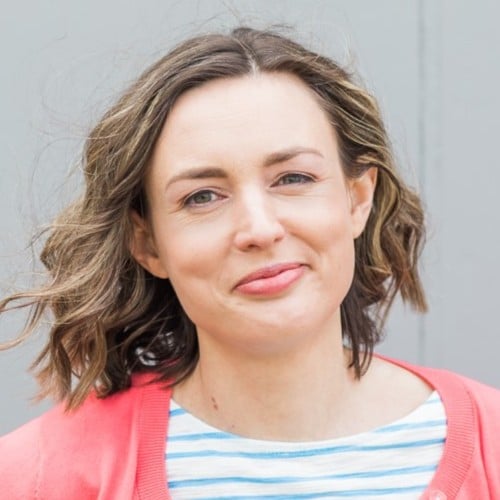A woman with wavy brown hair is smiling slightly. She is wearing a coral pink cardigan over a white and blue striped top, standing in front of a plain gray background.