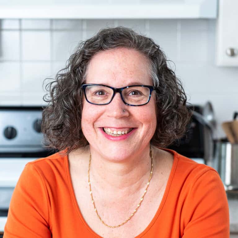 A person with glasses and curly hair is smiling while standing in front of a kitchen stove. They are wearing an orange top and a thin gold necklace. The background includes kitchenware and white tile walls.