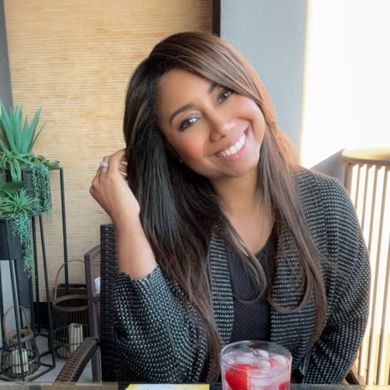 A woman with long brown hair smiles while sitting at a table indoors. She wears a black and white patterned jacket and holds her hair with one hand. A drink with ice and strawberries is on the table in front of her.