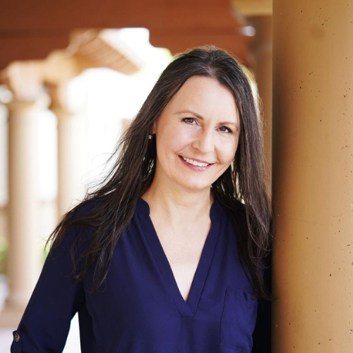 A woman with long dark hair, wearing a navy blue blouse, smiles while leaning against a pillar. The background features a series of columns and a blurred, sunlit walkway.