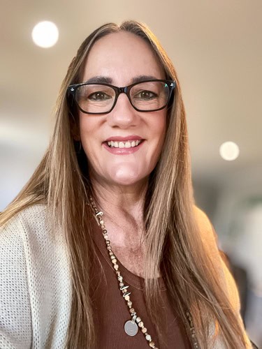 A smiling woman with long straight hair, wearing black glasses, a brown top, a beige cardigan, and a long necklace. The background is softly blurred, with indistinct lights.