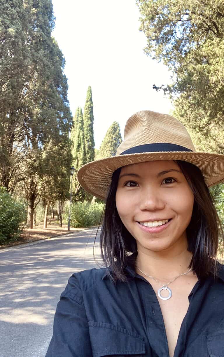 A woman smiles at the camera while taking a selfie outdoors. She wears a beige hat and a black shirt, and the background features a road lined with tall, lush trees under a bright sky.