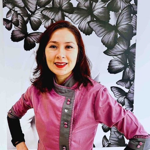 A woman in a purple chef’s jacket stands smiling with her hands on her hips in front of a black-and-white butterfly-patterned background.