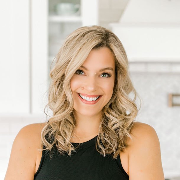 A young woman with wavy blonde hair smiles at the camera. She is wearing a black sleeveless top and standing in a bright, modern kitchen with white cabinets and a light-colored backsplash.