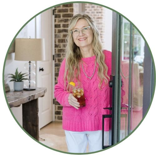 A smiling woman with long blonde hair and glasses, wearing a bright pink sweater and white pants, stands by an open door holding a glass of iced tea with a lemon slice. A lamp and potted plant are visible in the background.