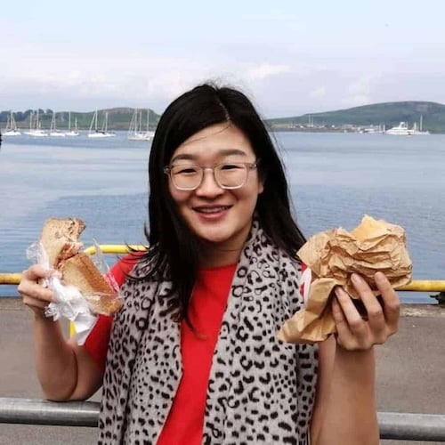 A smiling person with glasses holds a wrapped sandwich in each hand. They are standing in front of a scenic view of calm water and sailboats. The person is wearing a red shirt and a leopard print scarf.