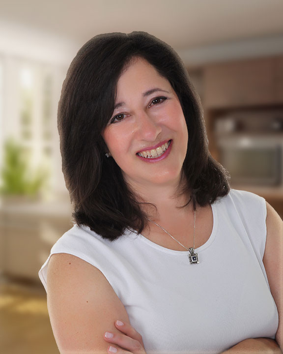 A woman with shoulder-length dark hair smiles warmly at the camera. She is wearing a white sleeveless top and a silver necklace with a pendant. Her arms are crossed. The background is a blurred indoor setting, which appears to be a kitchen.
