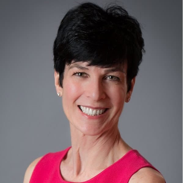 A woman with short black hair is smiling at the camera. She is wearing a bright pink sleeveless top and has small stud earrings. The background is plain and gray, putting focus on her cheerful expression.
