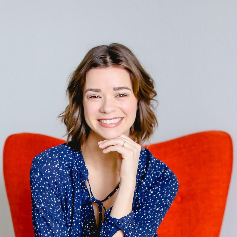A person with shoulder-length brown hair smiles at the camera, resting their chin on their hand. They are wearing a blue patterned blouse and are seated in front of a bright orange chair. The background is a plain light gray wall.