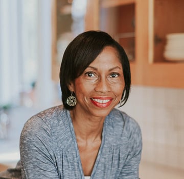 Smiling woman with shoulder-length black hair and red lipstick, wearing a gray top and round earrings, stands in a warmly lit kitchen.