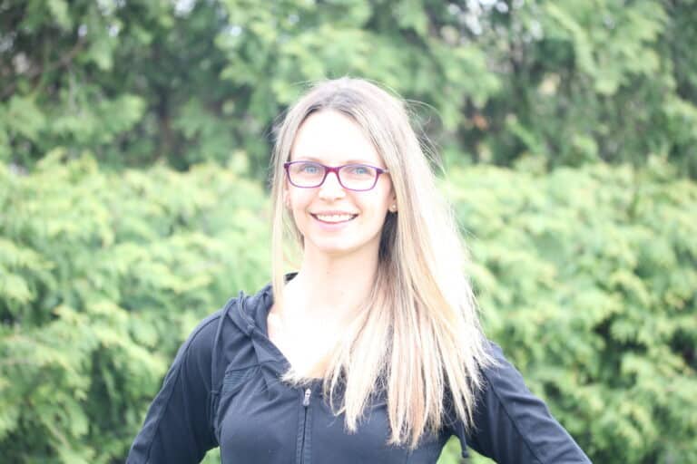 A woman with long blonde hair, wearing glasses and a black zip-up jacket, stands smiling in front of a lush green background of trees or bushes.