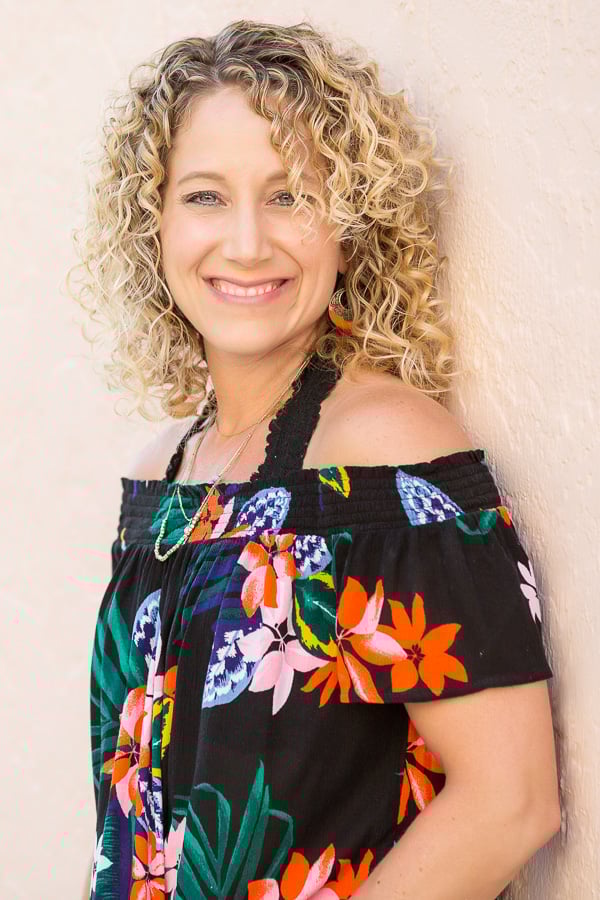 Woman with curly blond hair in a floral off-shoulder top, smiling and leaning against a light-colored wall.