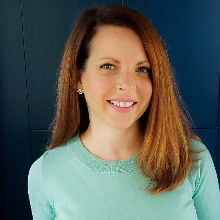 A woman with straight, shoulder-length auburn hair is smiling at the camera. She is wearing a light green top and gold earrings, standing in front of a dark blue background.