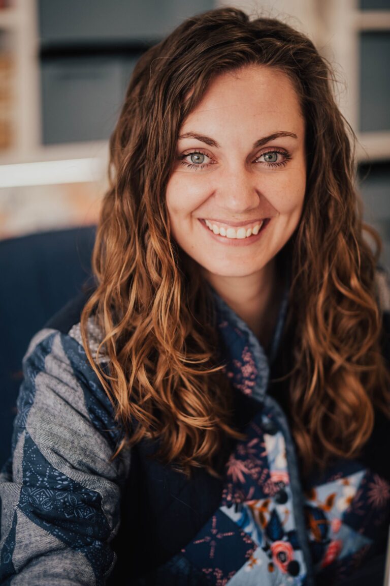 Smiling person with long wavy hair, wearing a patterned shirt with blue tones. They are indoors, sitting in front of a blurred background that includes shelving.
