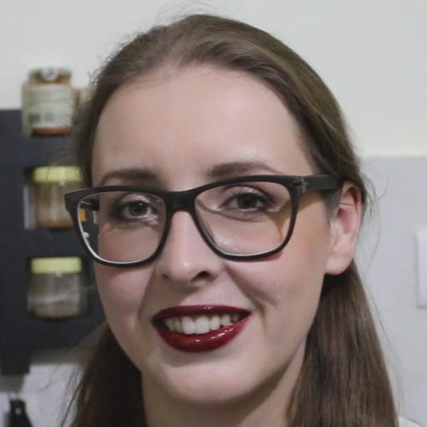 A woman with long brown hair is smiling. She is wearing glasses with dark frames and bold red lipstick. The background shows part of a kitchen with jars on a shelf and a light switch.