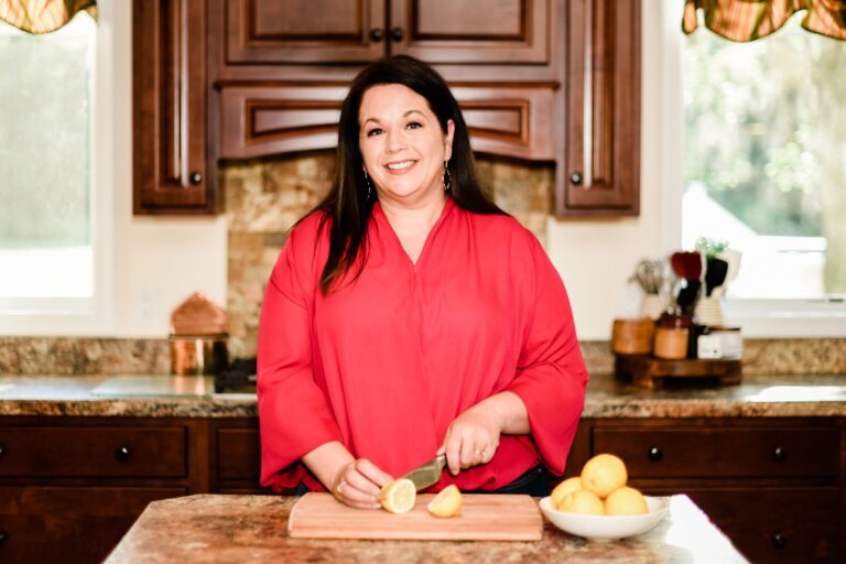 A person in a red blouse smiles while cutting lemons on a wooden board in a kitchen. The kitchen has brown cabinets, a stone backsplash, and a bowl of lemons on the counter. Bright light filters through windows in the background.