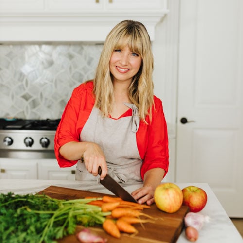 A person in a red top and apron smiles while chopping carrots on a wooden board in a kitchen. Apples, greens, and a shallot are nearby. A stove and white cabinets are in the background.
