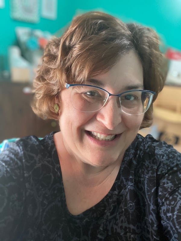 A person with short, wavy brown hair and glasses smiles at the camera. They're wearing a patterned black top. The background features a teal wall and blurred room elements.