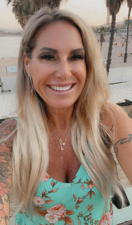 A woman with long blonde hair smiles at the camera. She is wearing a floral dress with a light blue background and gold jewelry. In the background, there is a beach with palm trees and a sunset sky.