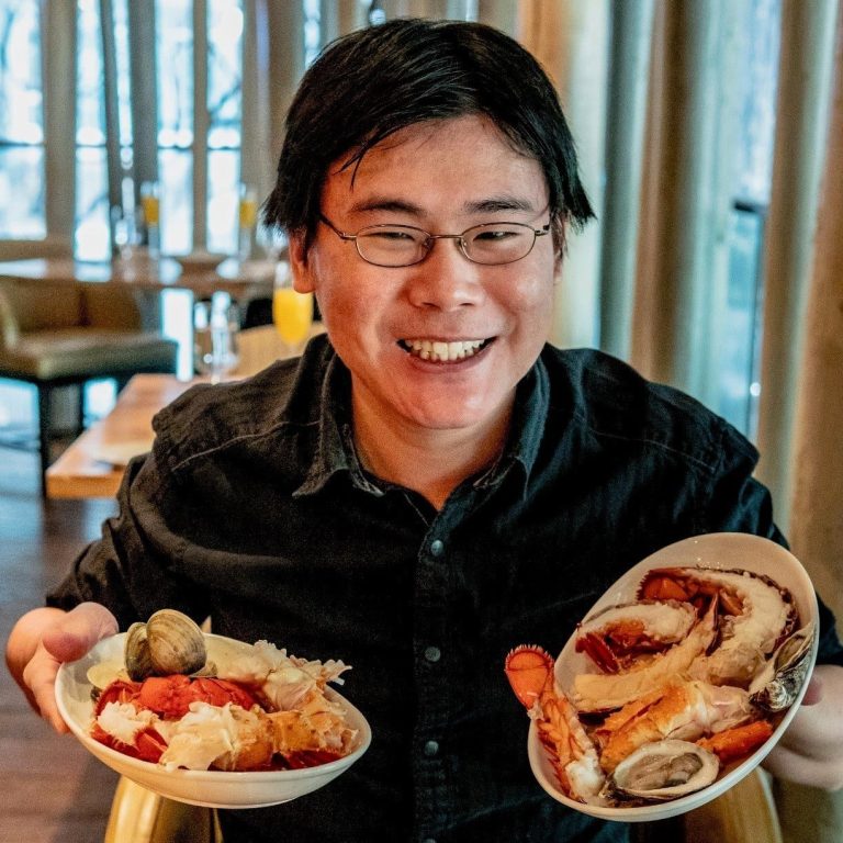 A smiling person with glasses and dark hair holds two bowls of assorted seafood, including lobster and clams, while seated in a restaurant with wooden chairs and large windows in the background.