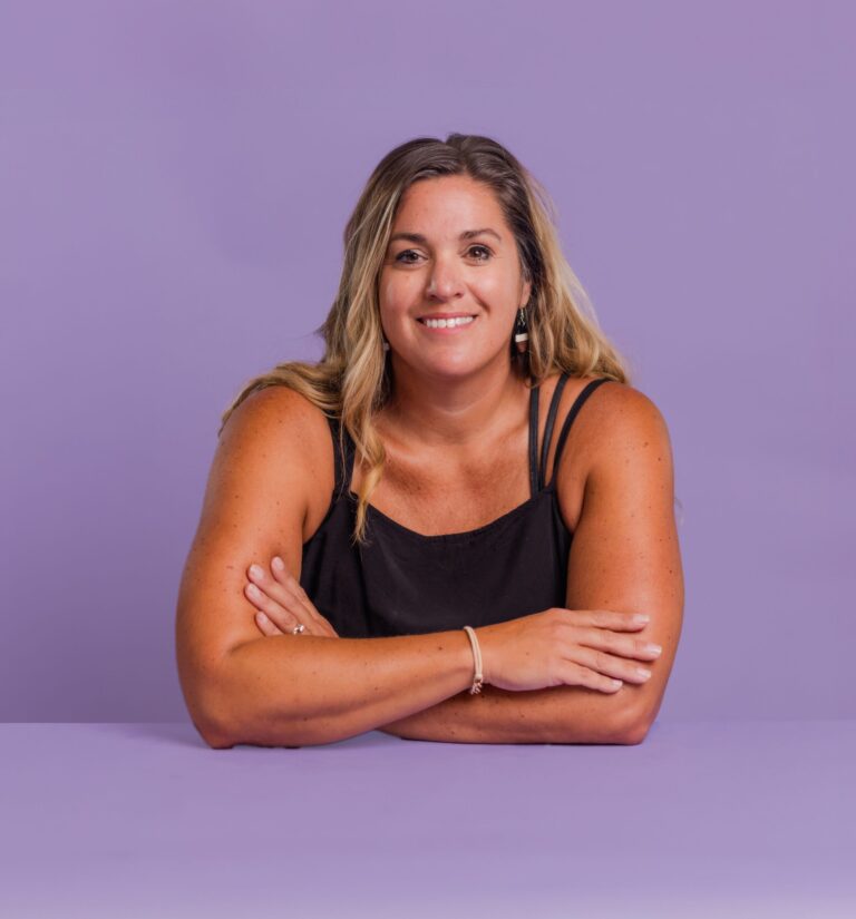 A woman with long hair, smiling, sits with her arms crossed on a purple surface. The background is also purple, and she is wearing a black sleeveless top.