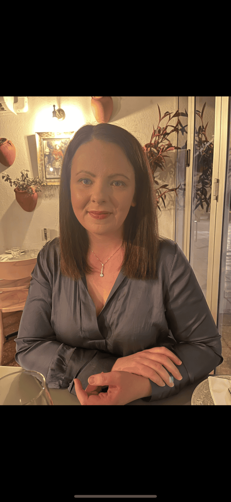 A woman with shoulder-length dark hair sits at a dining table, wearing a gray, long-sleeve blouse. She is smiling softly and has her arms casually crossed. The background features plants in pots on the wall and a soft warm light.