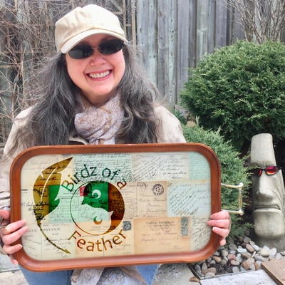 A person wearing sunglasses, a beige cap, and a scarf holds a decorative tray with "Birdz of a Feather" on it. They are smiling and standing outdoors near a plant and a stone face sculpture.