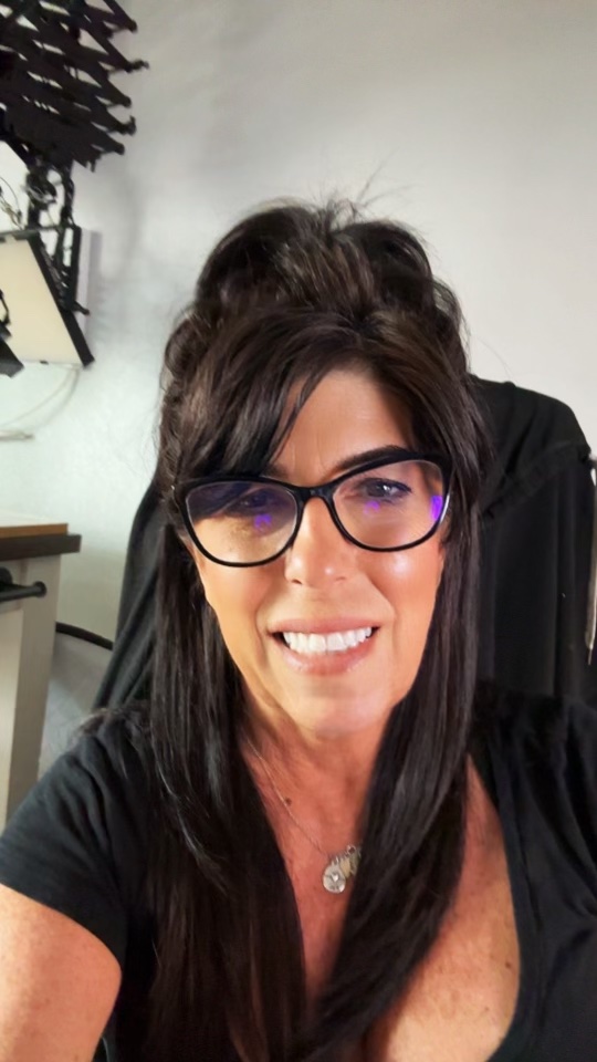 A woman with long dark hair and glasses smiles at the camera in a room. She is wearing a black top and a necklace with a round pendant. The background shows part of a desk with equipment.
