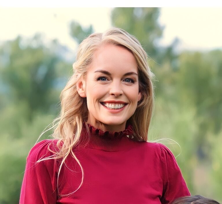 A person with long blonde hair smiles brightly while wearing a red high-neck top. The background is a natural setting with blurred greenery and trees.