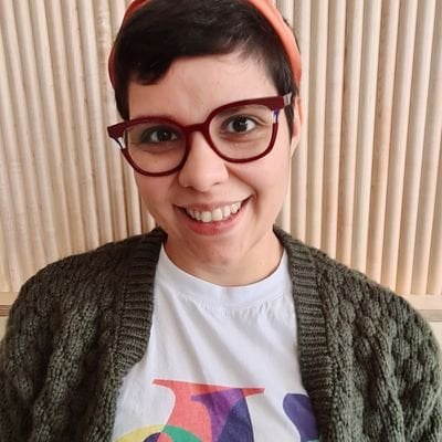 A person with short dark hair, wearing red glasses, an orange headband, a white T-shirt with colorful letters, and a green knit cardigan, smiling in front of a background with vertical wooden slats.