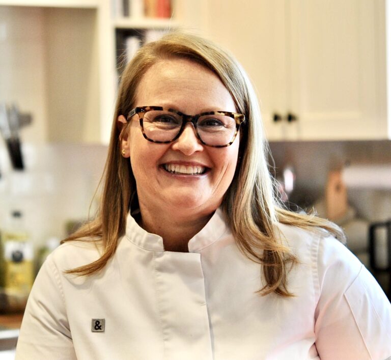A smiling woman wearing glasses and a white chef's coat stands in a kitchen with white cabinets. She has shoulder-length blonde hair and is looking directly at the camera, creating a warm and welcoming atmosphere.
