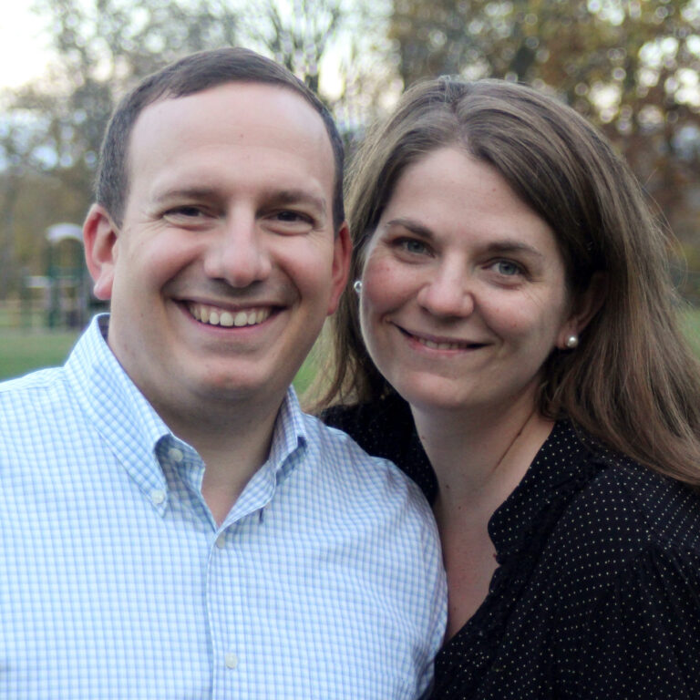 A man in a light blue button-down shirt and a woman in a dark blouse smile and pose closely together outdoors, with a blurred park and trees in the background.