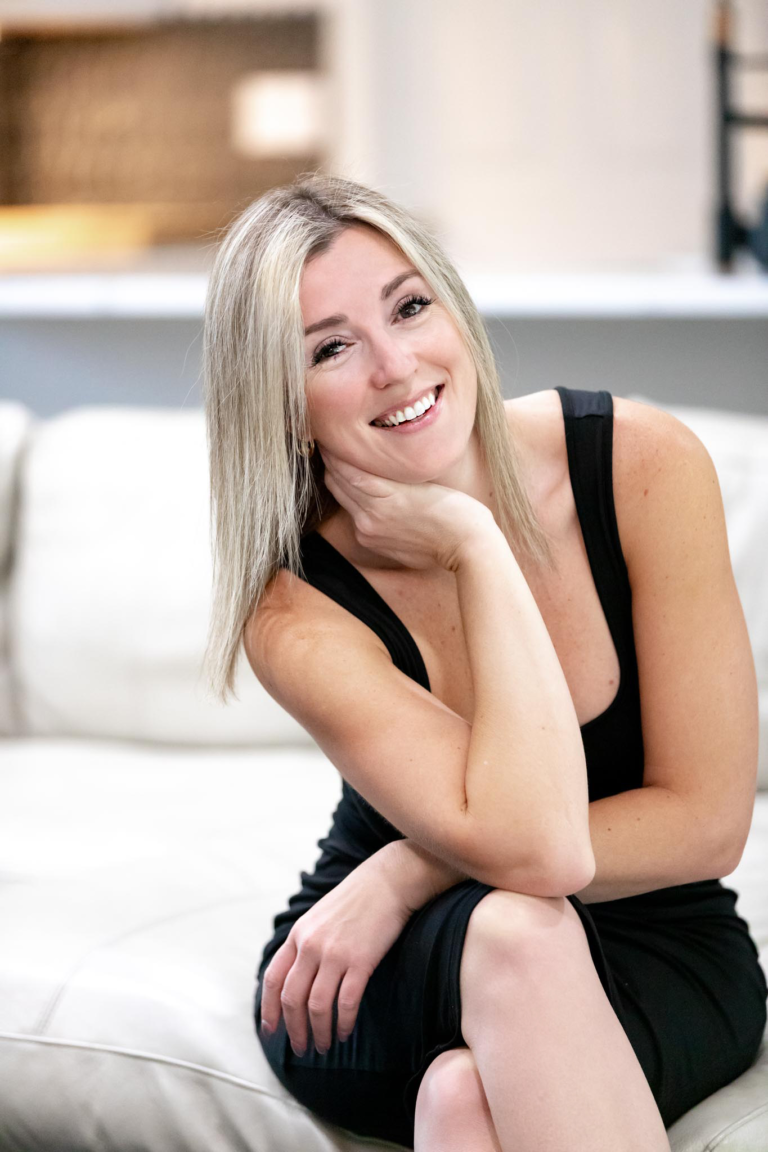 A smiling woman with blonde hair sits on a white couch, wearing a black sleeveless dress. Her chin rests on her hand, and she looks directly at the camera. The background is softly blurred, suggesting a cozy, modern interior.