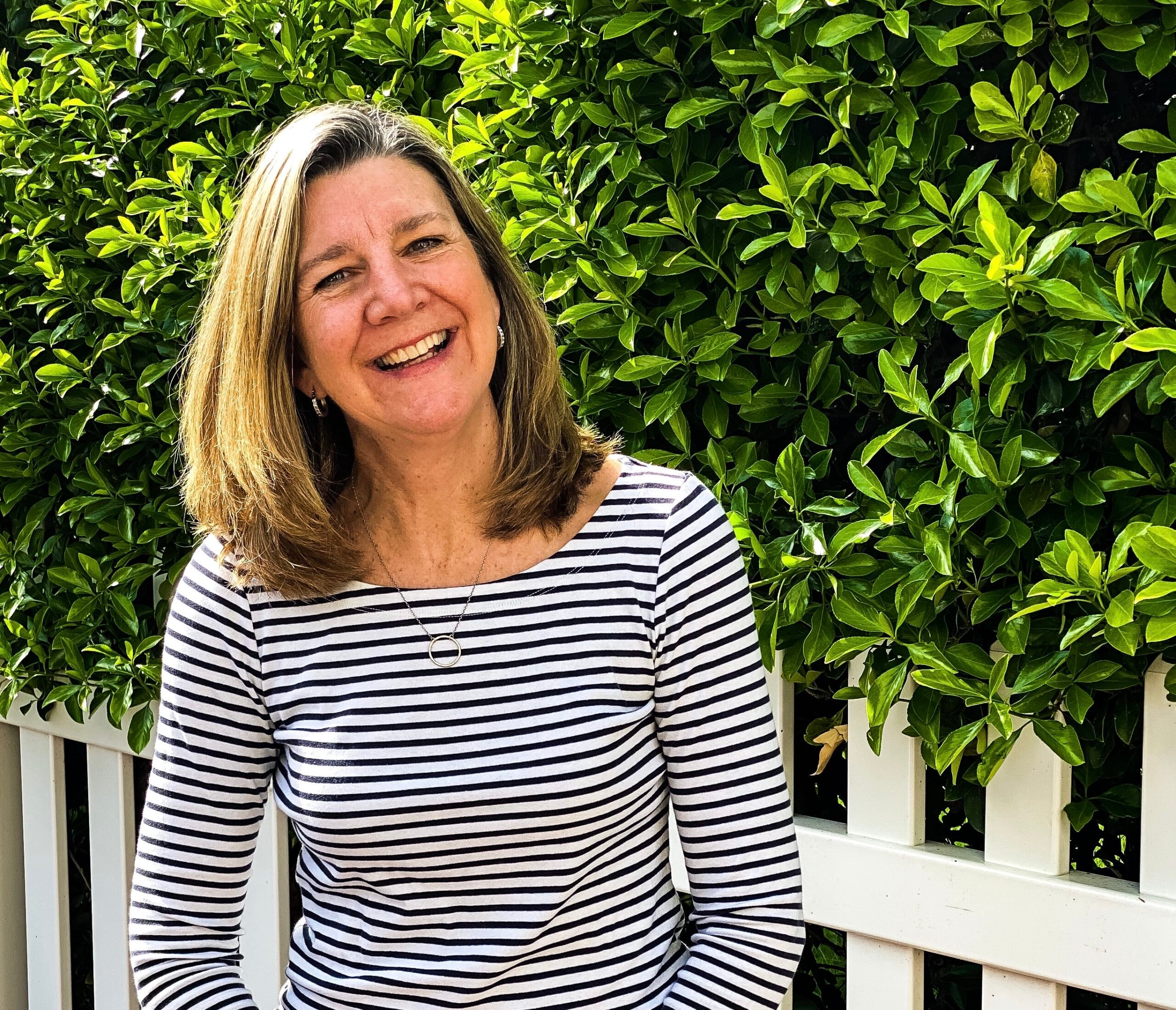 A woman with long hair smiles while sitting in front of a white fence. She is wearing a black and white striped shirt. Behind her, there is greenery with bright green leaves.