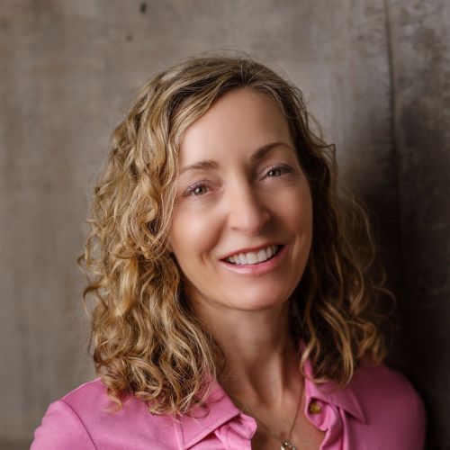 A smiling woman with shoulder-length curly blonde hair and light skin is wearing a pink collared shirt. She stands in front of a plain, textured gray wall, looking directly at the camera.