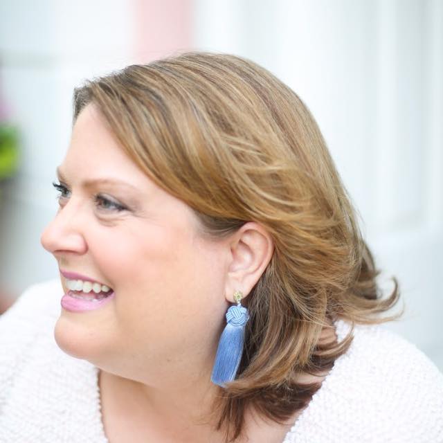 A woman with shoulder-length light brown hair smiles while looking to the left. She is wearing a light sweater and a long blue tassel earring. The background is softly blurred in pastel tones.