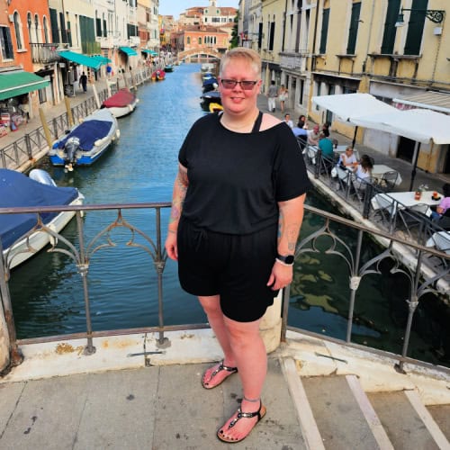 A person with short blonde hair and glasses, dressed in a black outfit, stands on a bridge over a canal with boats and colorful buildings in the background. People are dining at outdoor cafes along the canal. The scene is bright and sunny.