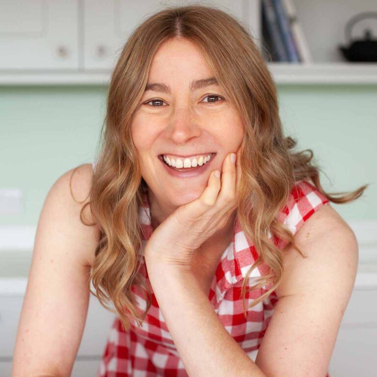 A person with wavy, light brown hair smiling warmly at the camera. They are wearing a red and white checkered sleeveless top and resting their chin on their hand. The background is a light-colored room with shelves.